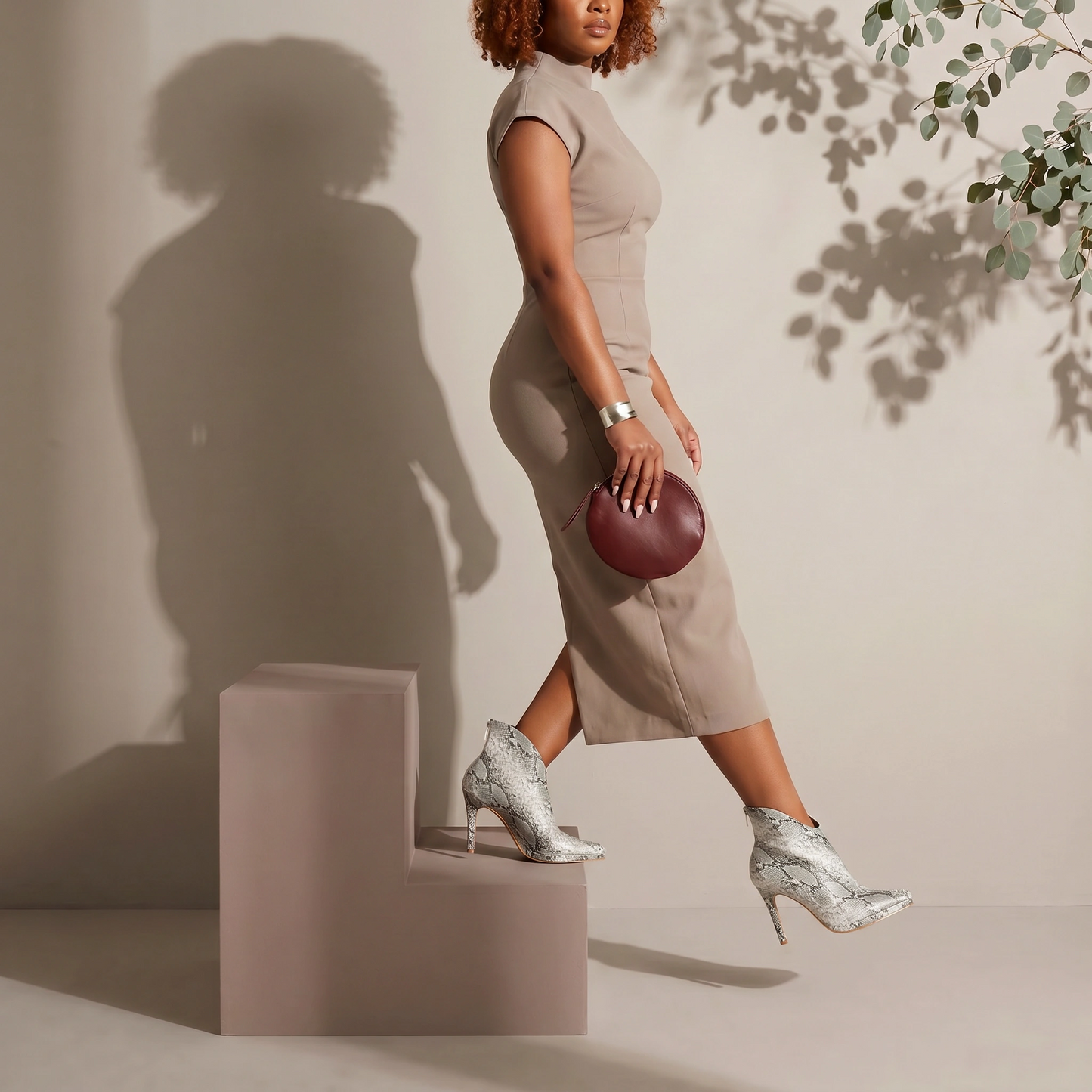 Woman in a beige dress and silver high heels standing on a pink block, holding a red clutch.