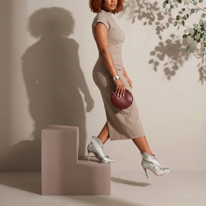 Woman in a beige dress and silver high heels standing on a pink block, holding a red clutch.